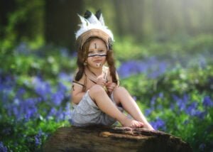 Child in whimsical costume sitting on a log amidst vibrant bluebells in Nottingham’s enchanted woods.