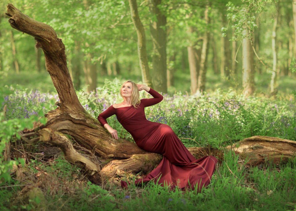 mum posing in a wood in a red maternity dress surrounder by bluebells flowers, Family session in Bluebells Wood in Nottingham