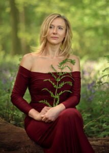 Serene woman in a burgundy dress sitting in a bluebell wood during her outdoor birthday photoshoot.