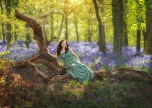 Woman in a green dress reclining on a fallen tree amidst bluebells in Nottingham's woodland during a spring photoshoot.