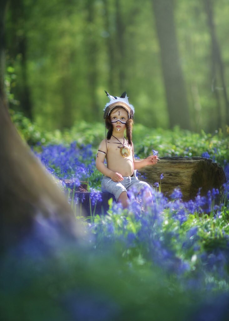 child sitting on a tree trunk in a middle of bluebell wood in Nottingham Forest wearing native indian headpiece during family and children photography