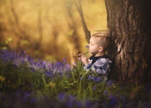 Thoughtful young boy sitting by a tree and holding a bluebell in Nottingham's woodland during a spring photoshoot.