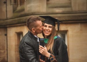 A proud partner kissing a graduate in academic regalia while holding her diploma on Nottingham University’s historic grounds.