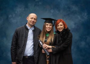 A graduate in an academic gown standing with her parents, holding a diploma during a Nottingham graduation photoshoot.