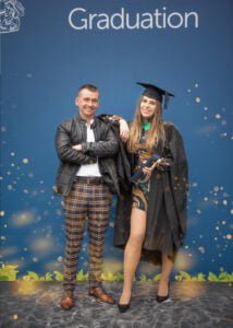 A graduate in regalia and her partner smiling at Nottingham Trent University’s official graduation backdrop.