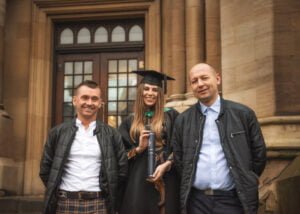 A graduate in academic regalia posing with two proud family members on the Nottingham University campus steps.