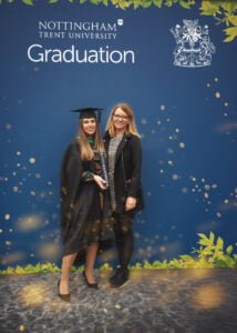 A graduate in regalia posing with her friend in front of Nottingham Trent University’s official graduation backdrop.