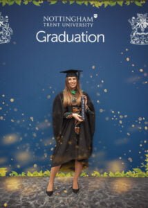 A graduate in an academic gown holding a diploma at Nottingham Trent University’s official graduation backdrop.