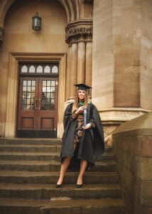 A graduate in an academic gown standing on the steps of a historic Nottingham University building with her diploma.