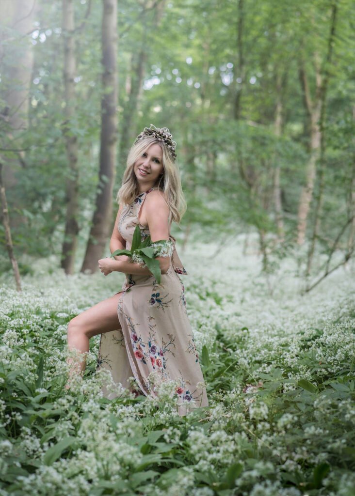 A woman adorned with a floral crown smiles gently in a lush Nottingham forest, surrounded by wild garlic flowers.