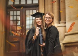 A graduate and her friend smiling on the Nottingham University campus, surrounded by autumn leaves and holding a diploma.