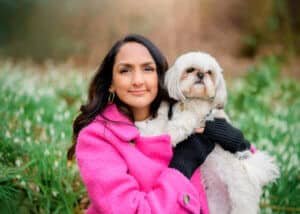 A woman in a vibrant pink coat lovingly cradles her white Shih Tzu in a field of green, sharing a moment of connection and love.