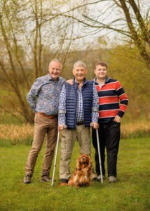 Three generations of family stand together outdoors with their golden spaniel sitting loyally at their feet, embodying a legacy of companionship and love.