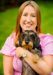 A joyful woman in a pink dress shares a loving embrace with her adorable wire-haired dachshund, capturing the essence of companionship in Nottingham's lush greenery.
