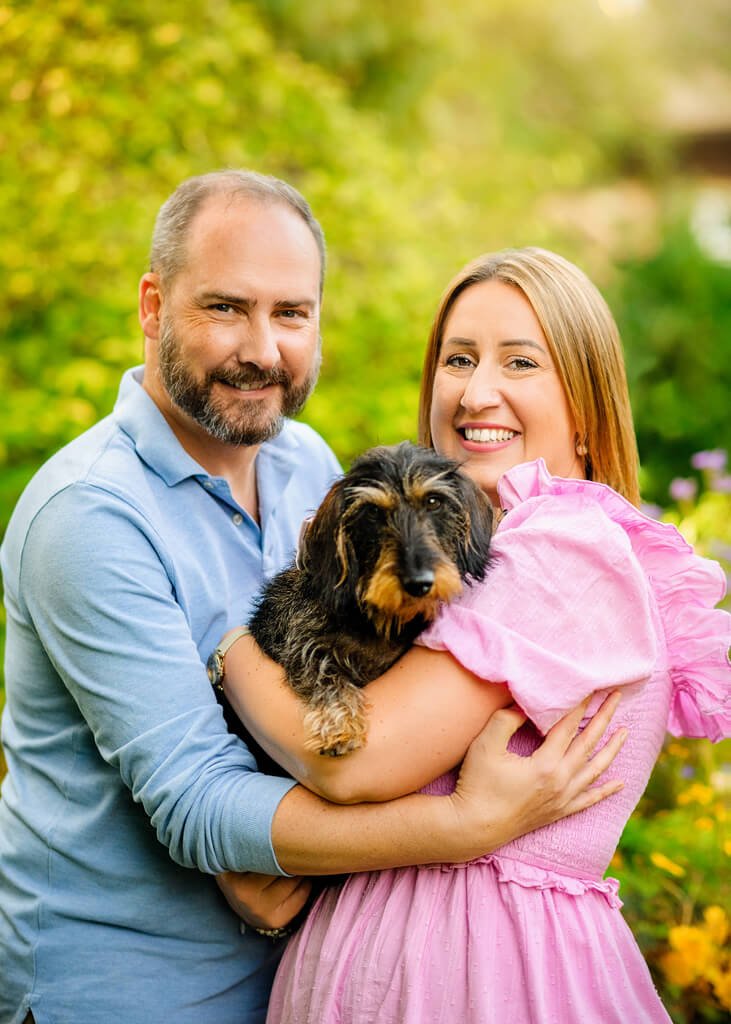 A couple in a tender embrace smiles radiantly, holding their beloved dachshund, as they are enveloped in the soft glow of a sunlit garden.
