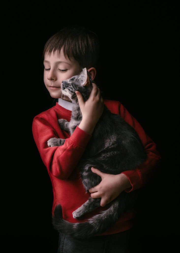 A young boy, eyes closed in contentment, gently cradles a grey and white cat in his arms against a dark background, a silent bond of companionship.