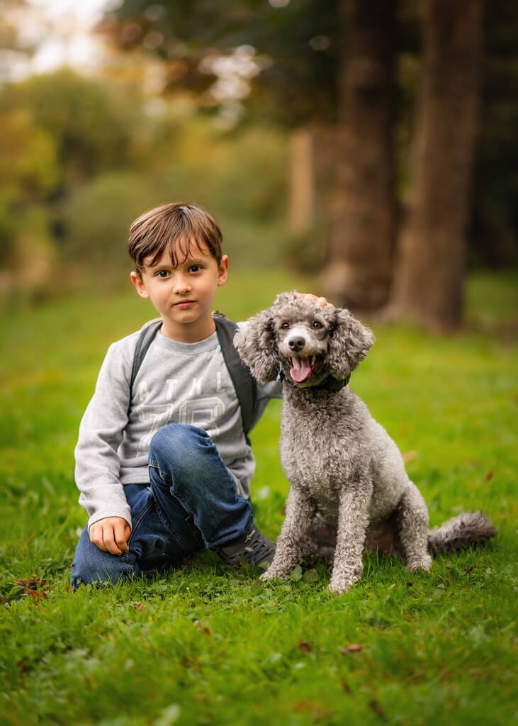 A young boy kneels beside his joyful grey poodle in the lush greenery of a Nottingham park, showcasing the bond of friendship and love in a natural portrait session.