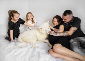 A family enjoys a playful moment with their white dog on a cozy rug, with laughter and gentle pets shared between parents and daughters.