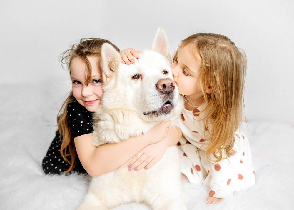 Two young girls share a tender moment with their fluffy white dog, hugging and kissing him on a white fur rug, exuding affection and joy.