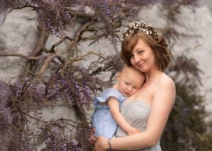 Mother embracing her young child among purple wisteria flowers during an outdoor photoshoot.