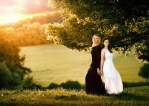Mother and teenage daughter standing under a tree at sunset, wearing black and white dresses.