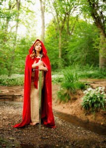 Woman in red cape holding a sword in a forest creek, professional portrait.