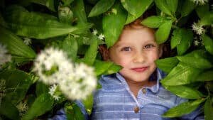 Smiling young child peeking through lush green leaves with delicate white flowers, eyes sparkling with joy.