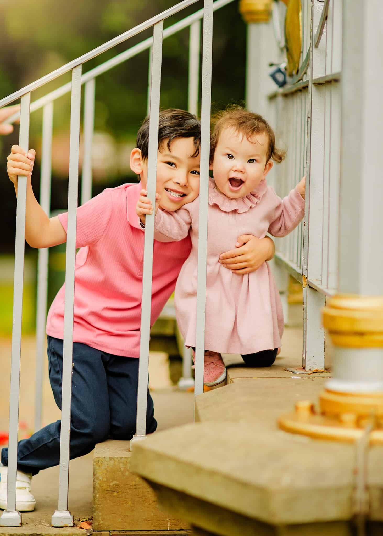 Two joyful children, a boy and a girl in coordinating pink outfits, playfully peek through white metal railings on a wooden porch, radiating happiness and sibling love.