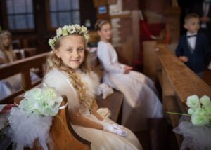 Girl with a floral crown sitting in a church pew during her First Holy Communion ceremony.