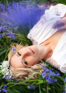 Woman in a lacy white dress reclines amidst bluebells, her gaze captivating in the soft light of her outdoor birthday photoshoot.