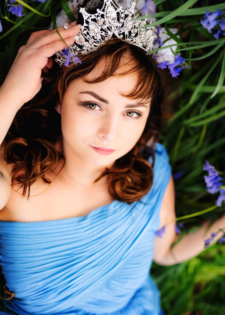 Close-up of a woman in a blue gown and tiara during an outdoor birthday photoshoot in a field of bluebells.
