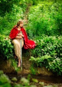 A woman in a golden dress and red cloak sits regally beside a brook, her outdoor birthday photoshoot evoking a fantasy realm.