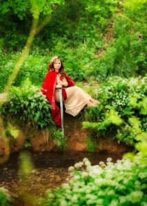 A regal woman in a red cloak and tiara sits by a woodland stream, her posture poised and reflective amid the tranquil backdrop of Nottingham's natural splendor.