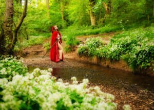 A woman clad in red, wielding a sword, stands empowered amidst the wild garlic by a Nottingham stream, her portrait a testament to strength and the spirit of adventure.
