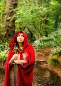 Enveloped in a red cloak and holding a sword, a woman stands in Nottingham's lush woods, her gaze lifted as if seeking guidance from the ancient whispers of the trees.
