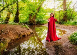 In the lush, verdant surroundings of Nottingham's woodlands, a woman in a resplendent red cloak stands by a stream, her contemplative gaze adding a touch of mystique to the serene environment.