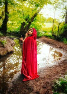 A woman stands by a woodland stream in Nottingham, draped in a scarlet cloak, her expression one of enchanting curiosity, as she holds a sword, symbolizing strength and elegance.