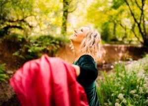 A woman in joyful abandon stretches her arm out, her face turned towards the sun filtering through the leaves, embodying the spirit of freedom in a Nottingham forest setting.