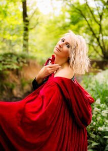 Woman draped in a vibrant red cape looking upwards in a lush forest setting, professional portrait photography.