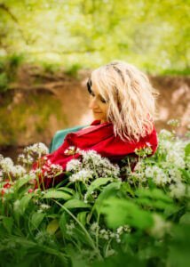 A woman, wrapped in a vibrant red shawl, sits amidst the lush greenery, her sideward glance capturing a moment of quiet contemplation in Nottingham's vibrant woodlands.