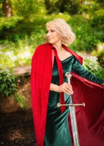 Woman in green dress and red cape holding a sword in a professional portrait.