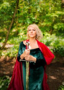 A woman in a green velvet dress and red cloak stands with a sword, exuding nobility during her outdoor birthday photoshoot.