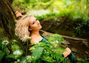 A serene woman in a green dress lounges by a woodland stream, her birthday photoshoot harmonizing with the lush forest backdrop.