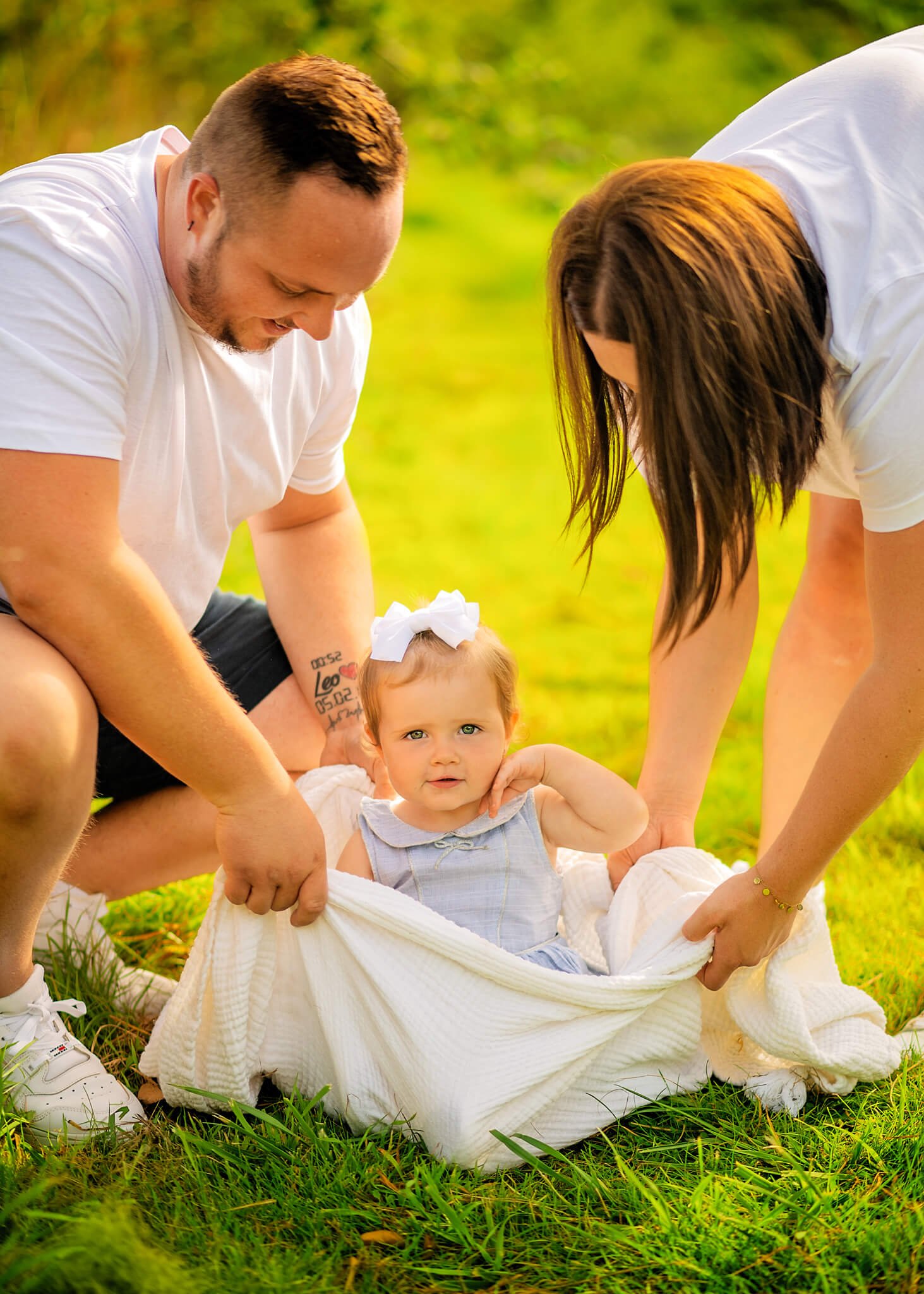 Toddler girl with a bow in her hair sitting on a blanket, flanked by her doting parents in a sunlit grassy field during a family photoshoot for kids.