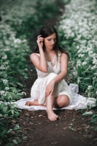 A young woman in a delicate white dress sits contemplatively on a forest path, surrounded by a blanket of white flowers, embodying a moment of peaceful reflection in a Nottingham women's portrait.