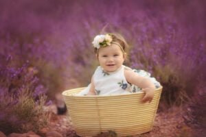 A gleeful baby sits in a woven basket amidst a sea of lavender, her smile as captivating as the vibrant purple that envelops her.