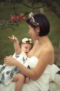A mother breastfeeding her baby under an apple tree, with the baby playfully reaching out and wearing a floral headband.