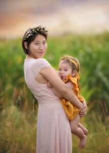 Mother in a soft pink dress holding her baby, who is breastfeeding, surrounded by a lush green field.