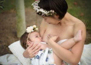 Mother breastfeeding her baby under a tree, both wearing floral crowns, showcasing a serene breastfeeding photography moment.