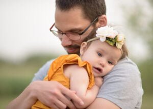 A father tenderly holding his baby girl in an orange dress with a floral headband, capturing a moment of paternal love.